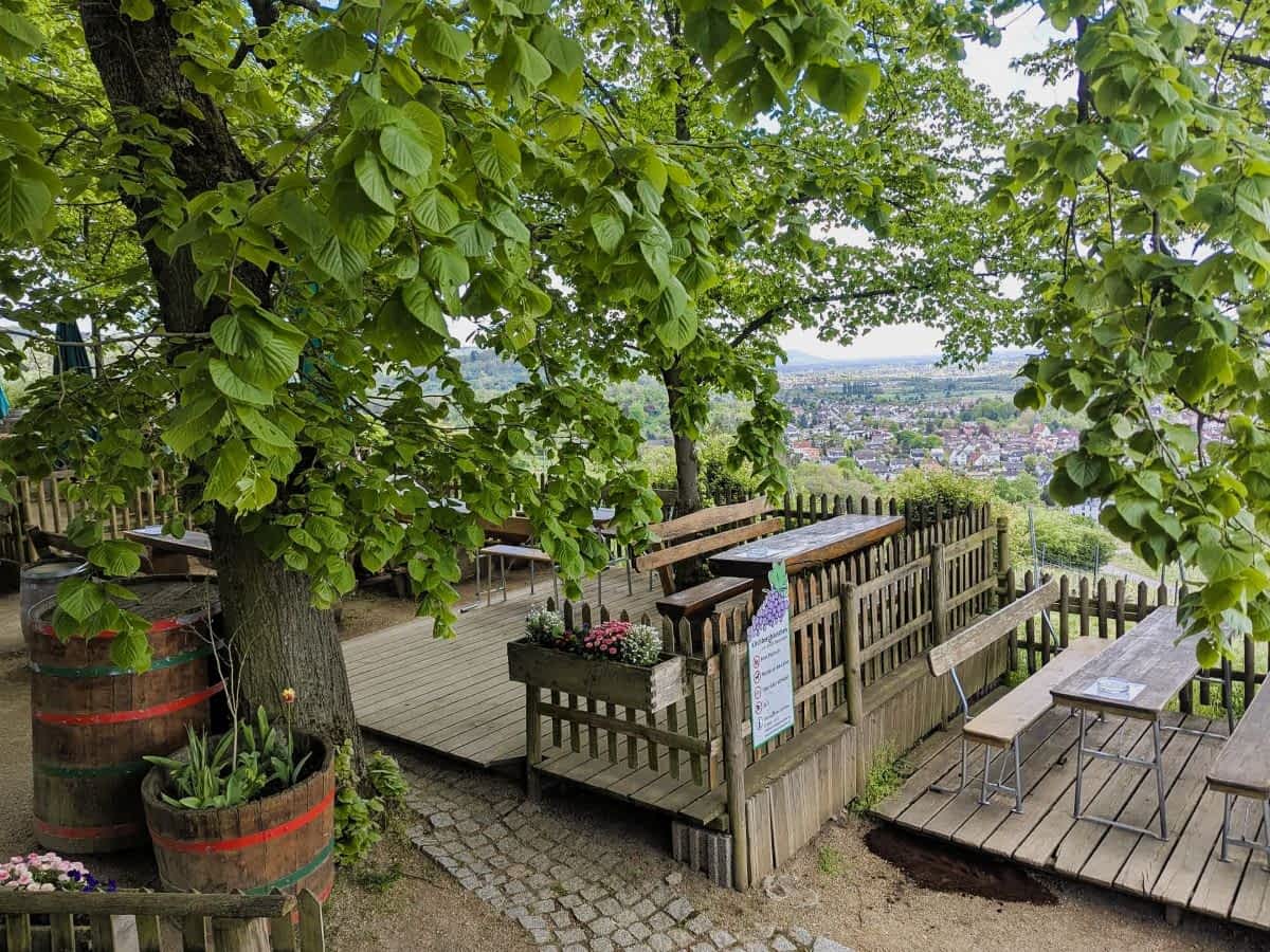  Rustikale Holzterrasse mit Biertischen und Bänken, umgeben von großen, grünen Bäumen und bepflanzten Weinfässern, mit Blick auf eine Kleinstadt im Tal.